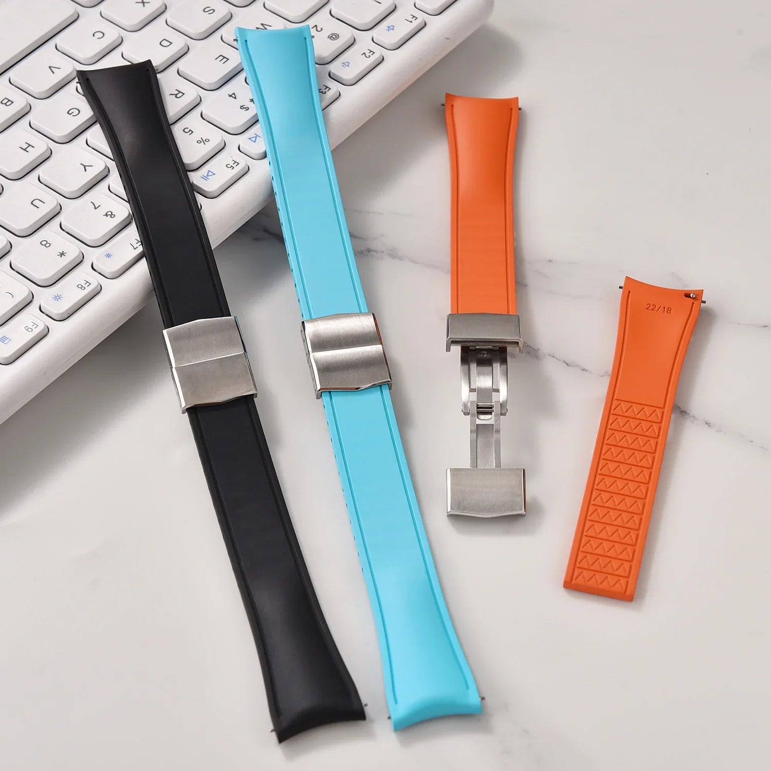Bracelets de montre en silicone noir, bleu et orange avec fermoirs en métal, posés sur un bureau blanc près d’un clavier, accessoires pour montres de luxe misterchrono.com.